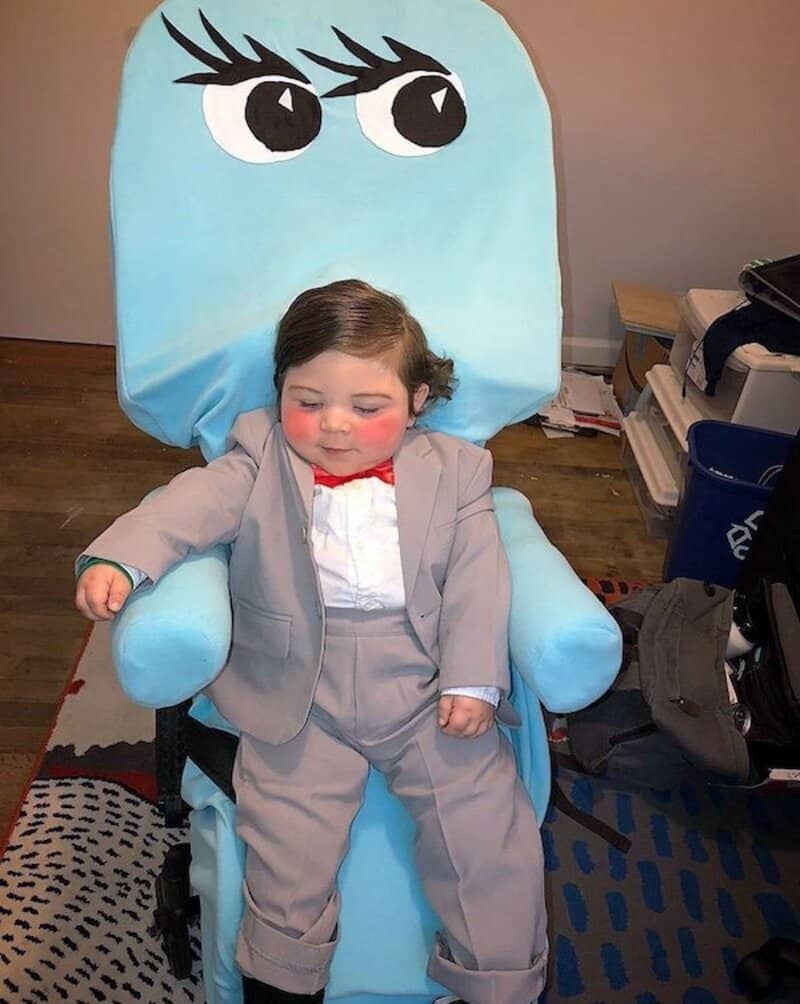 Child in light gray suit sitting on a light blue chair with large cartoon eyes, white shirt, red bow.
