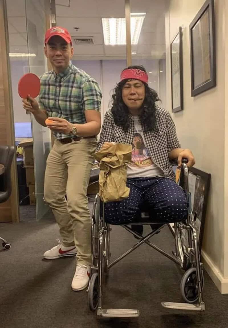 Two people indoors, one in a wheelchair with a pink headband, the other standing with a red cap, in.