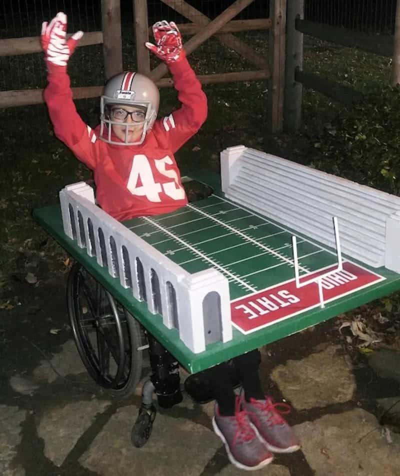 Person in a wheelchair wearing a football jersey number 45 with a cardboard stadium costume and goal.