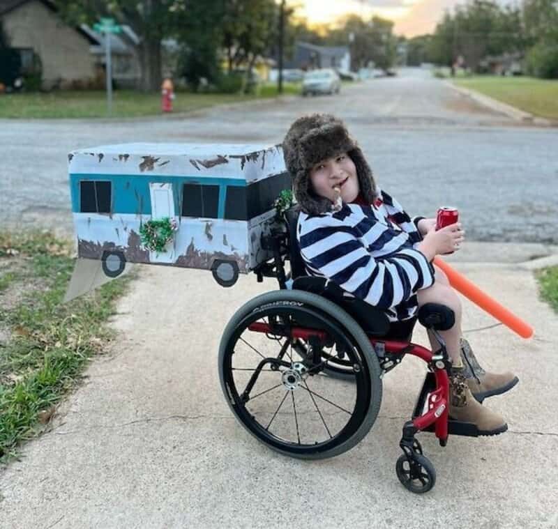 Person in a wheelchair with a cardboard bus costume on a suburban sidewalk.
