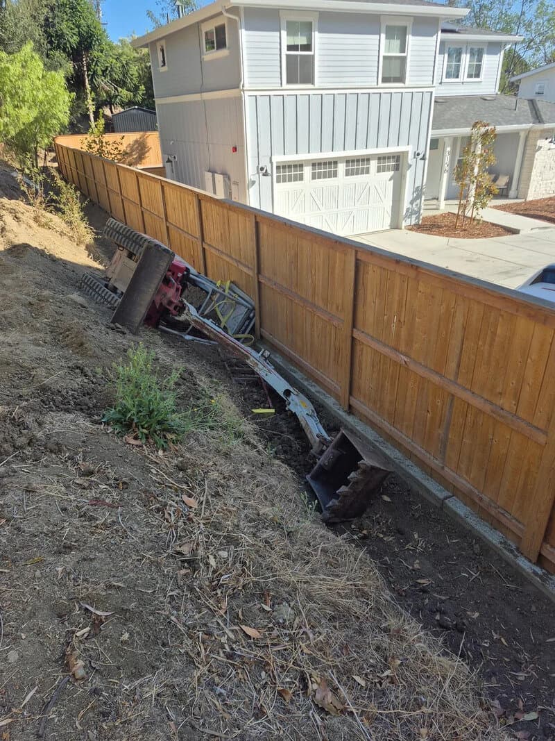 Construction vehicle tipped over on a slope next to a wooden fence and house.