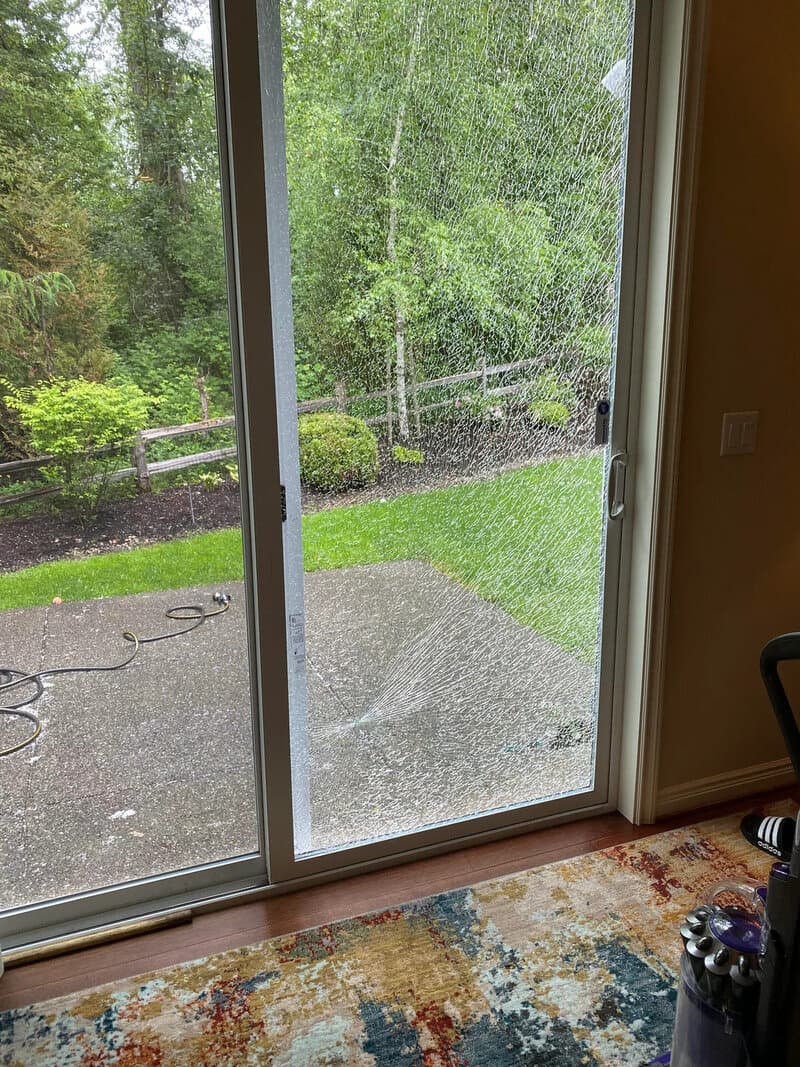 Rain pouring on sliding glass door with view of wet patio and green trees outside.