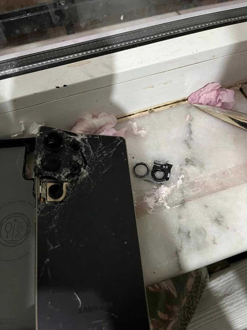 Broken phone with shattered screen and detached camera lens on a windowsill.