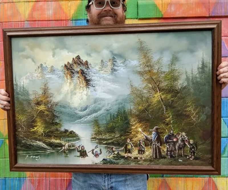 Landscape painting of mountains, trees, and people by a river, held by a man with sunglasses.