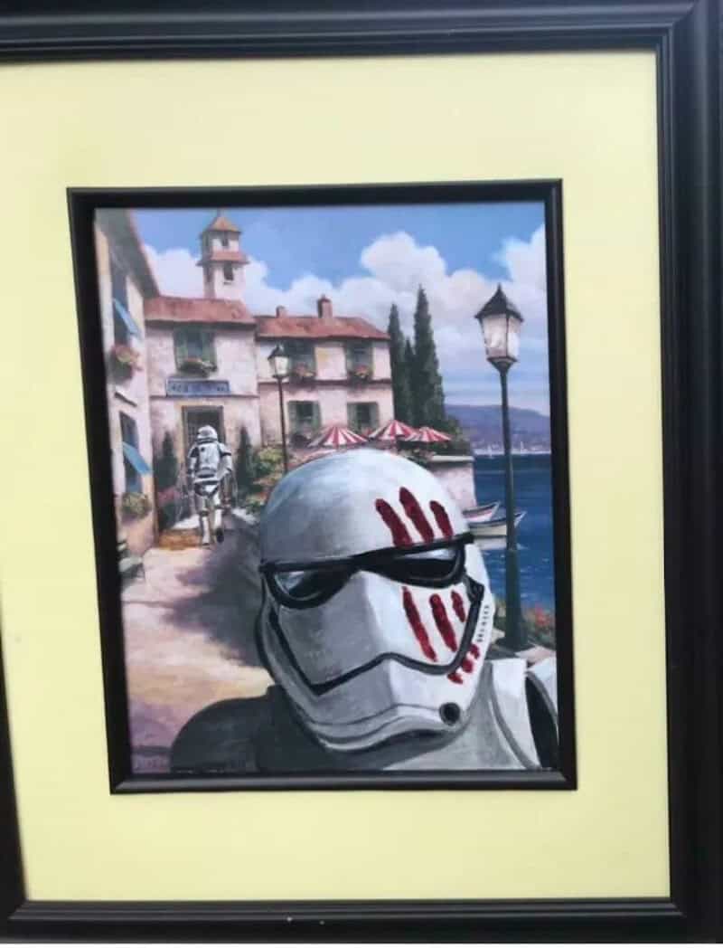 Painting of a stormtrooper helmet with red markings, set against a seaside village background.