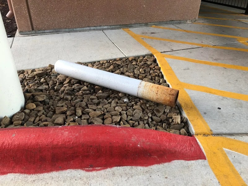 Rusty metal pipe on gravel next to yellow parking lines and sidewalk.