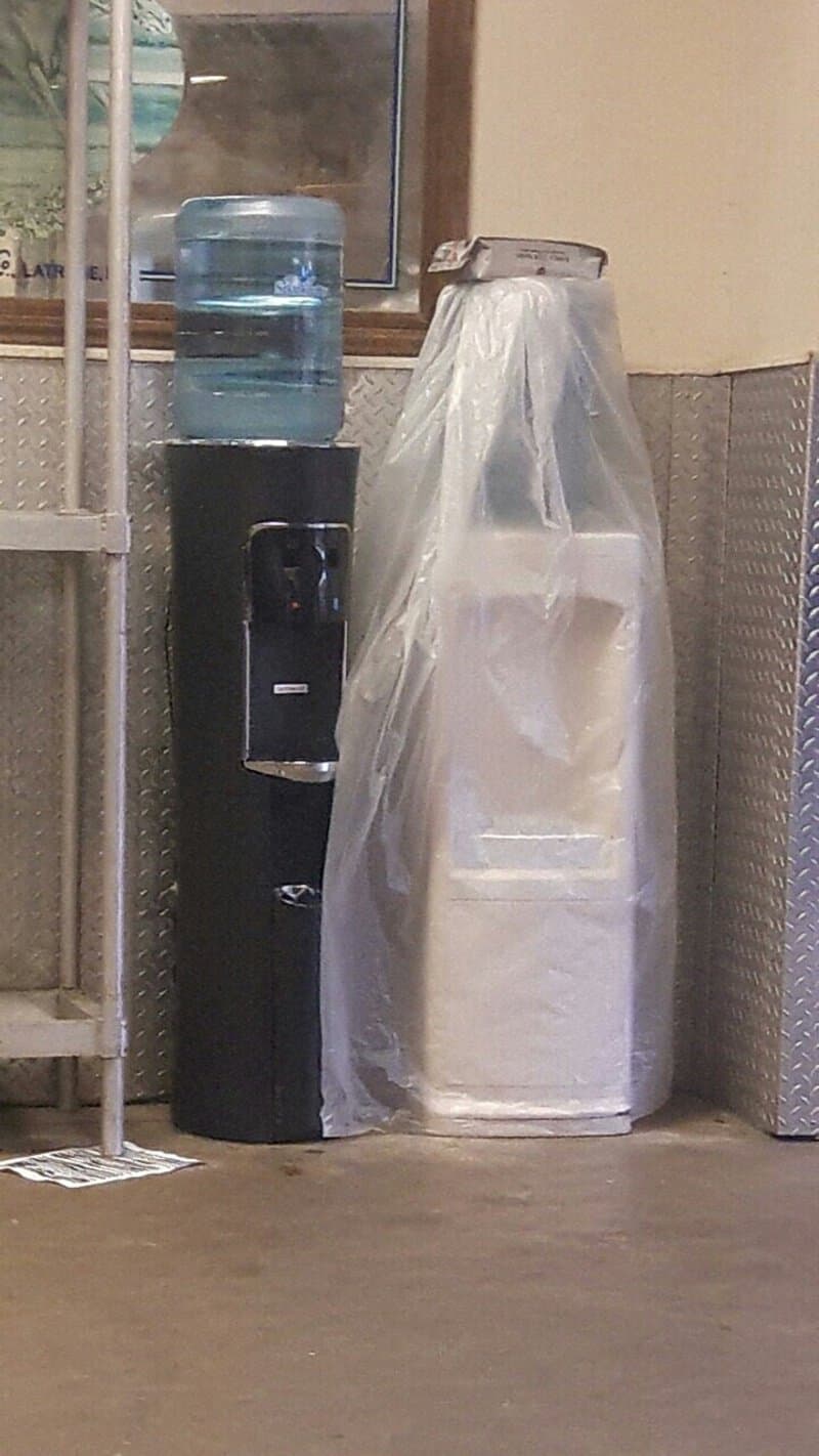 Water cooler and a large white bag with a handle, both against a wall.