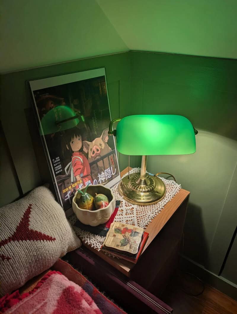 Green desk lamp on wooden nightstand with a bowl of fruit, a book, and a framed anime poster.