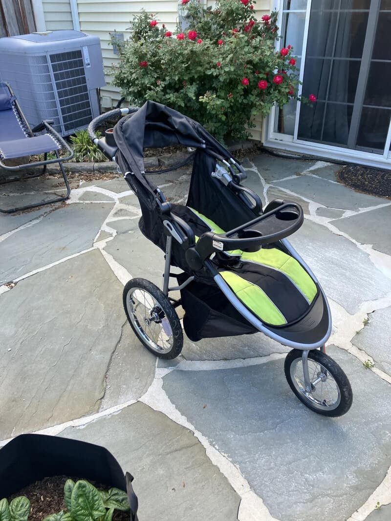 Black and green baby stroller on stone patio near house with flowers and air conditioning unit.