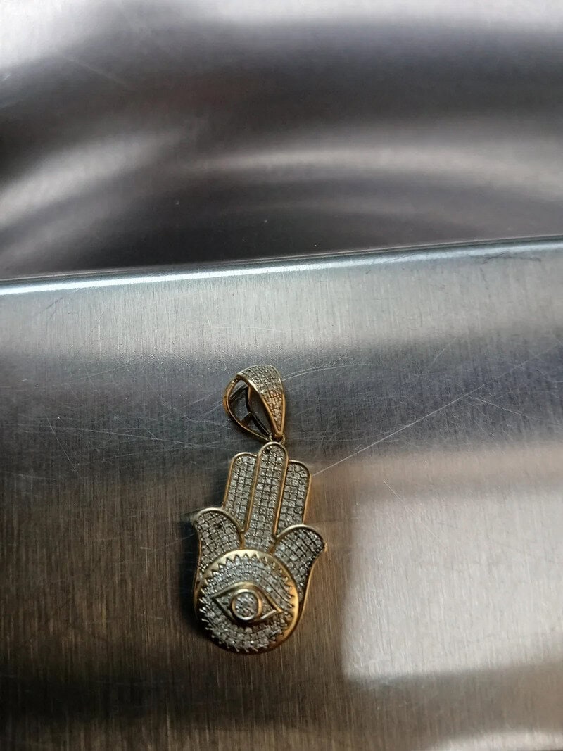 Gold pendant with eye and hand design, encrusted with small diamonds, on a metallic surface.