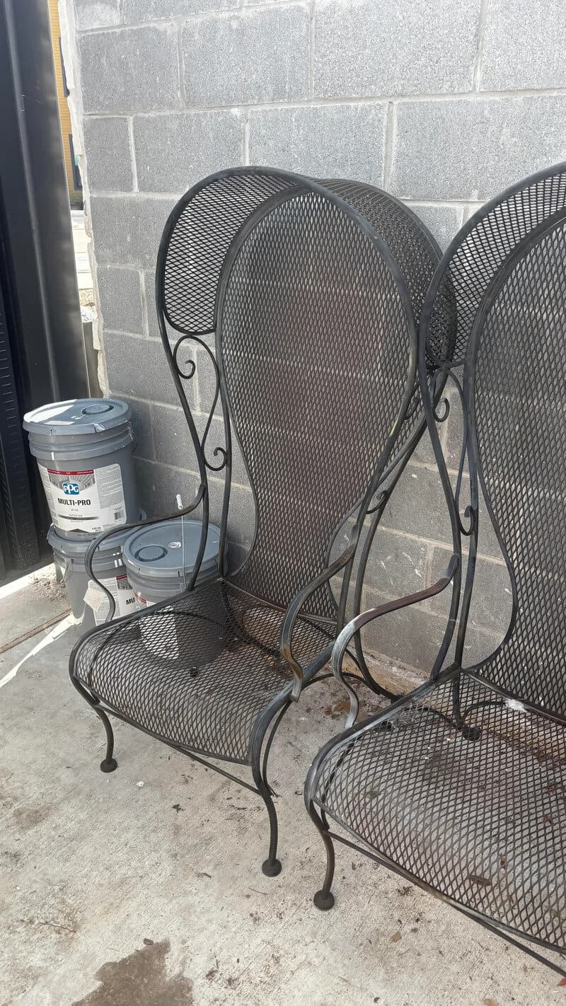 Two black metal outdoor chairs with mesh seats and backs, placed outside near a gray brick wall.