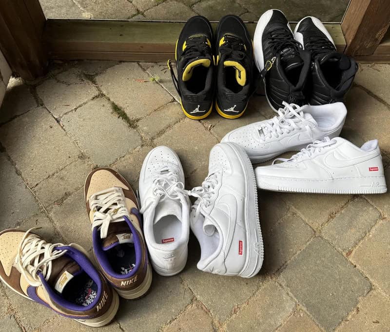 Collection of sneakers on a brick floor, some are white, black, purple, and beige.