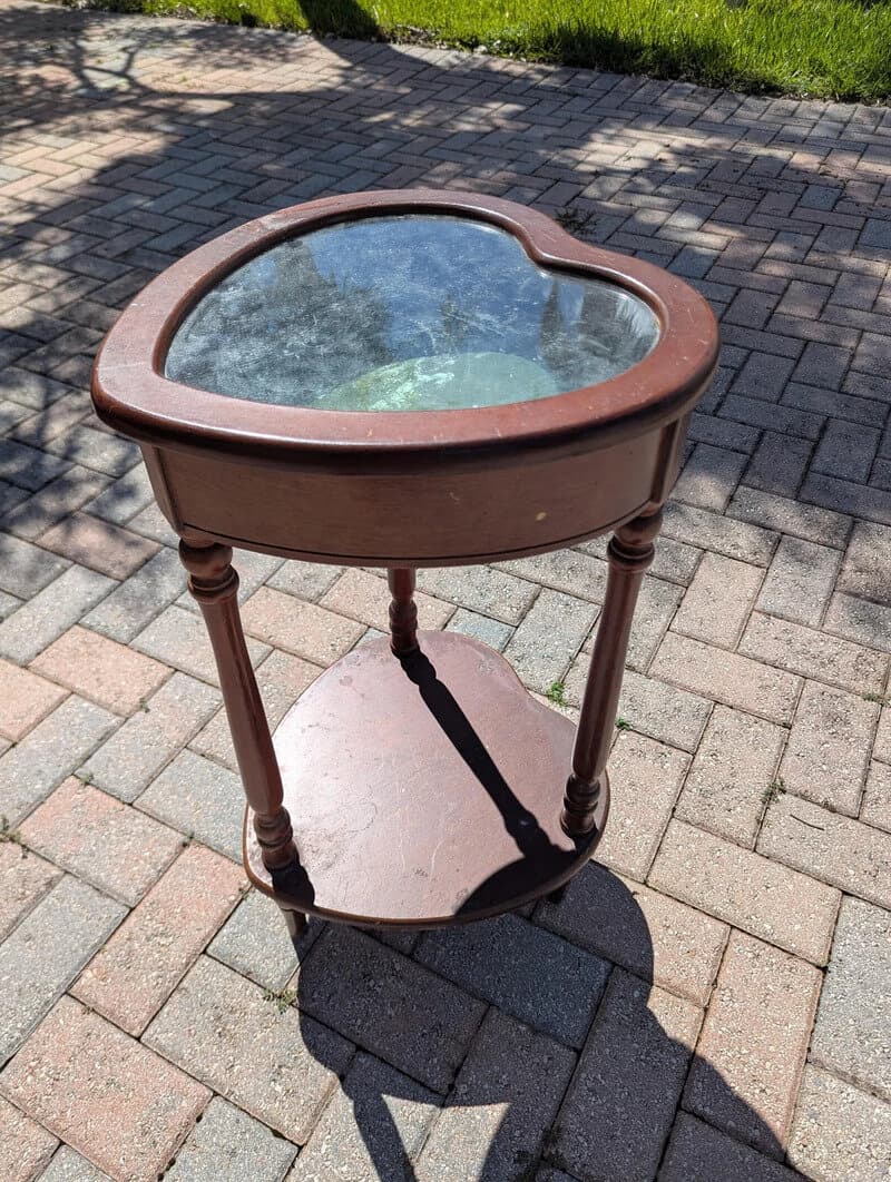 Heart-shaped glass-top table with wooden legs on a brick sidewalk.