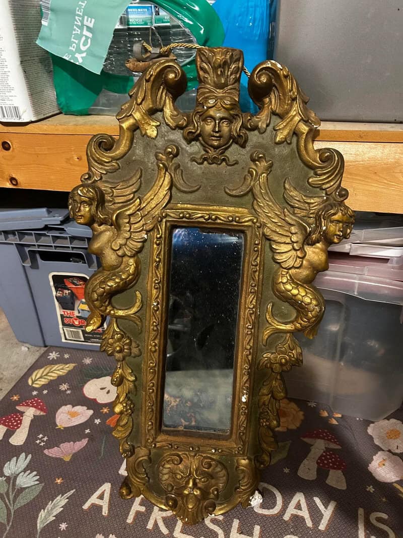 Ornate gold-framed mirror with cherubs and a face at the top, leaning against a storage area.
