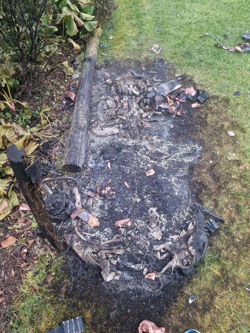 Charred remains of a burnt object on grass with debris and blackened area.