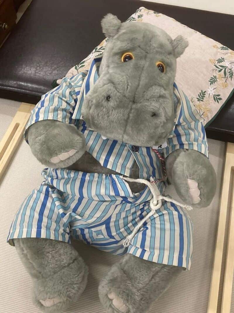 Gray hippopotamus plush toy in blue striped dress with white belt sitting on a bed.