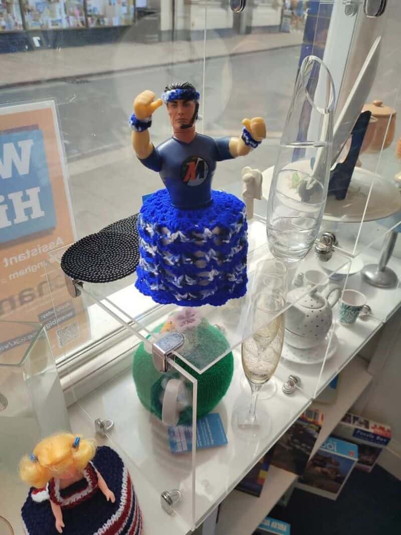 Action figure wearing blue outfit with raised fists atop a blue crocheted skirt in a shop display.