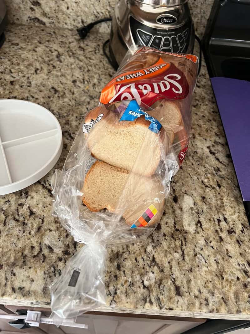 Sliced bread in a plastic bag on a kitchen counter with a bread machine nearby.