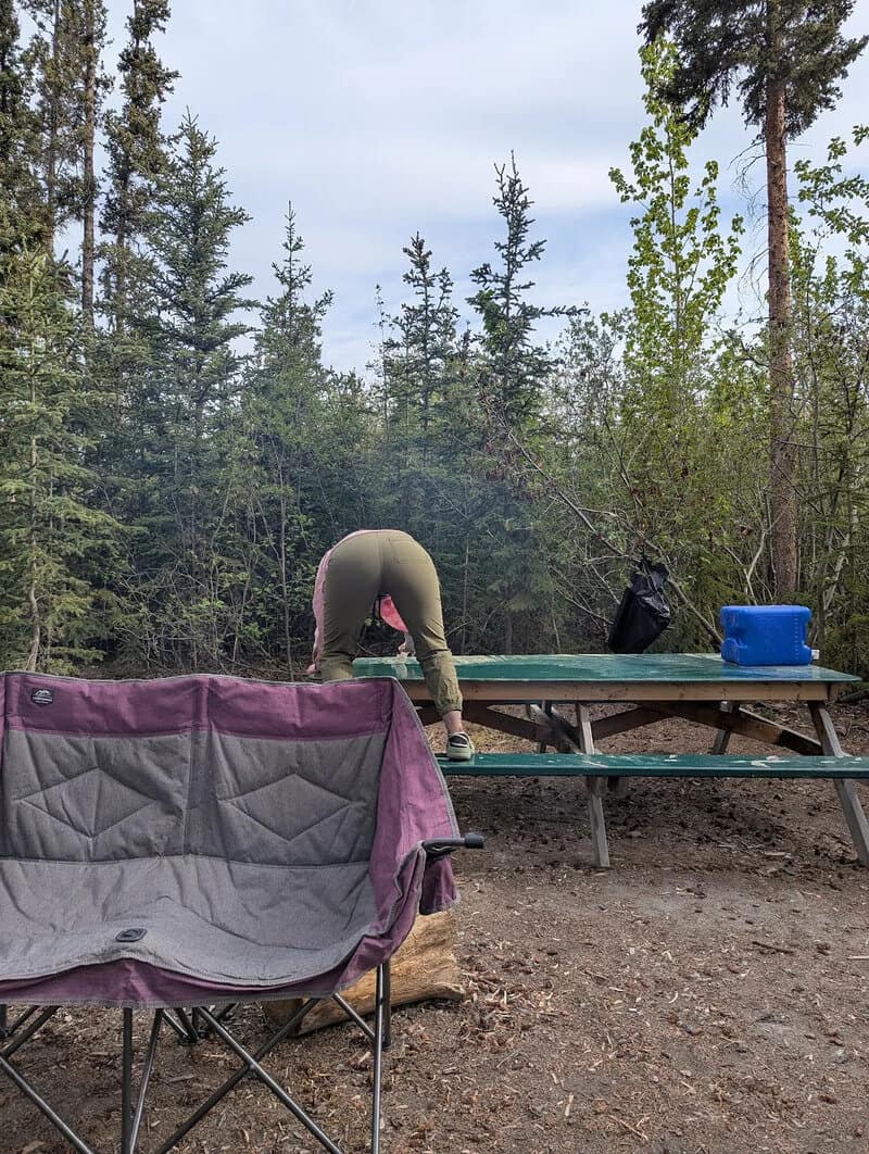 Person bending over at a picnic table in a forest setting.